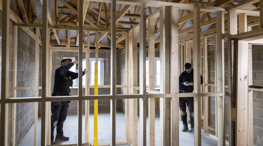 Wide shot of two male builders working on a property in the North East of England. They are  wearing work clothing, working together to manage the project. The frame of the new build is half complete around them. One of the men is attaching the frame together while the other is using his mobile phone.

Videos are available similar to this scenario.